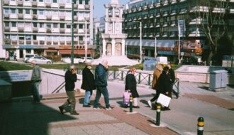 City street view with people walking near a prominent clock tower on a sunny day.