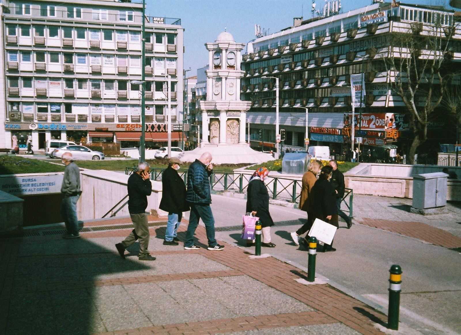 City street view with people walking near a prominent clock tower on a sunny day.