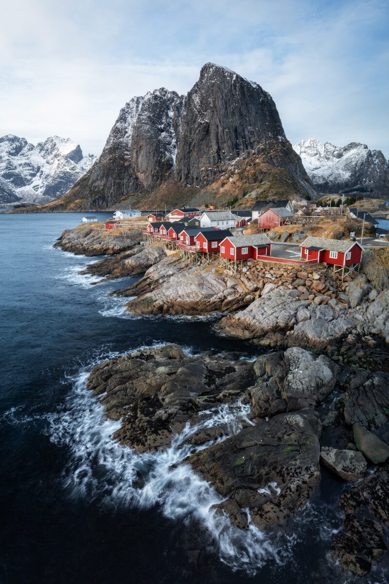 Red houses line a rocky coast with snow-capped mountains.