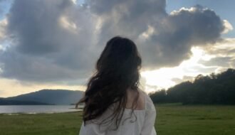 A woman in a white dress stands in a field, gazing at the dramatic clouds at sunset over the lake.