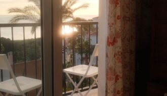 A tranquil sunrise view from a bedroom, overlooking a balcony with palm trees and ocean.