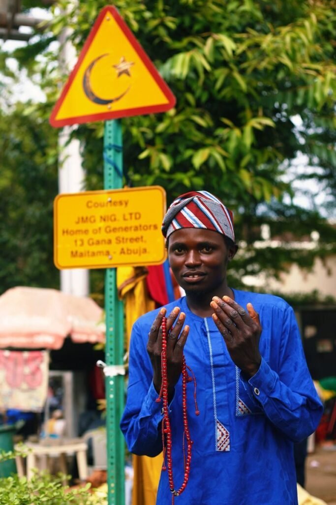 A man in traditional attire holding prayer beads, standing outdoors in Abuja, Nigeria, near signage.