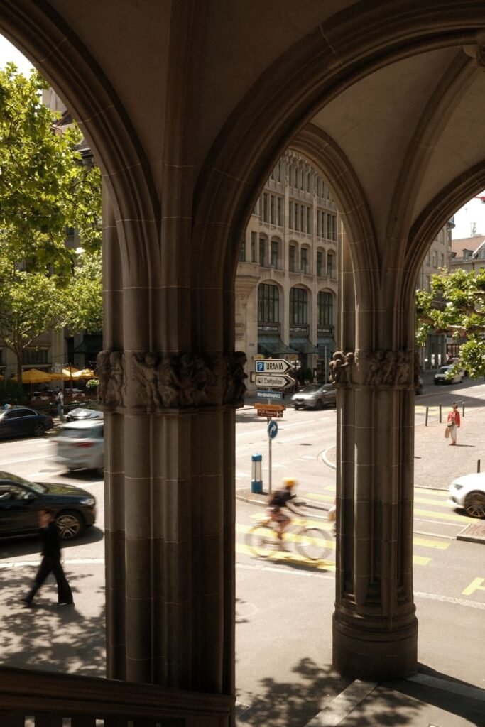A view through stone arches onto a busy street in Zurich, Switzerland, featuring blurred cyclists and cars.