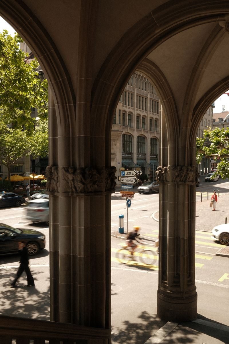 A view through stone arches onto a busy street in Zurich, Switzerland, featuring blurred cyclists and cars.