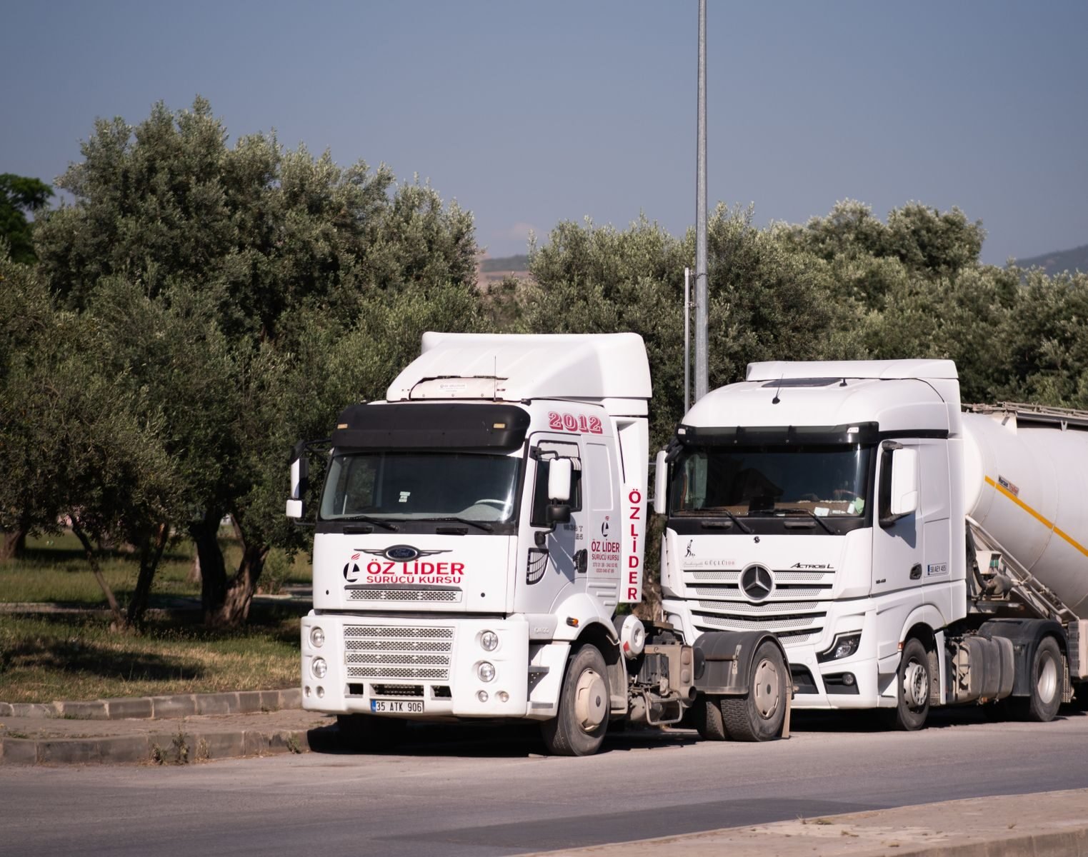 Two trucks parked on a road near olive trees in İzmir, Türkiye.