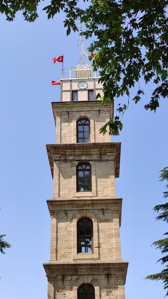 Stone clock tower with red flags and bright blue sky, framed by green leaves.