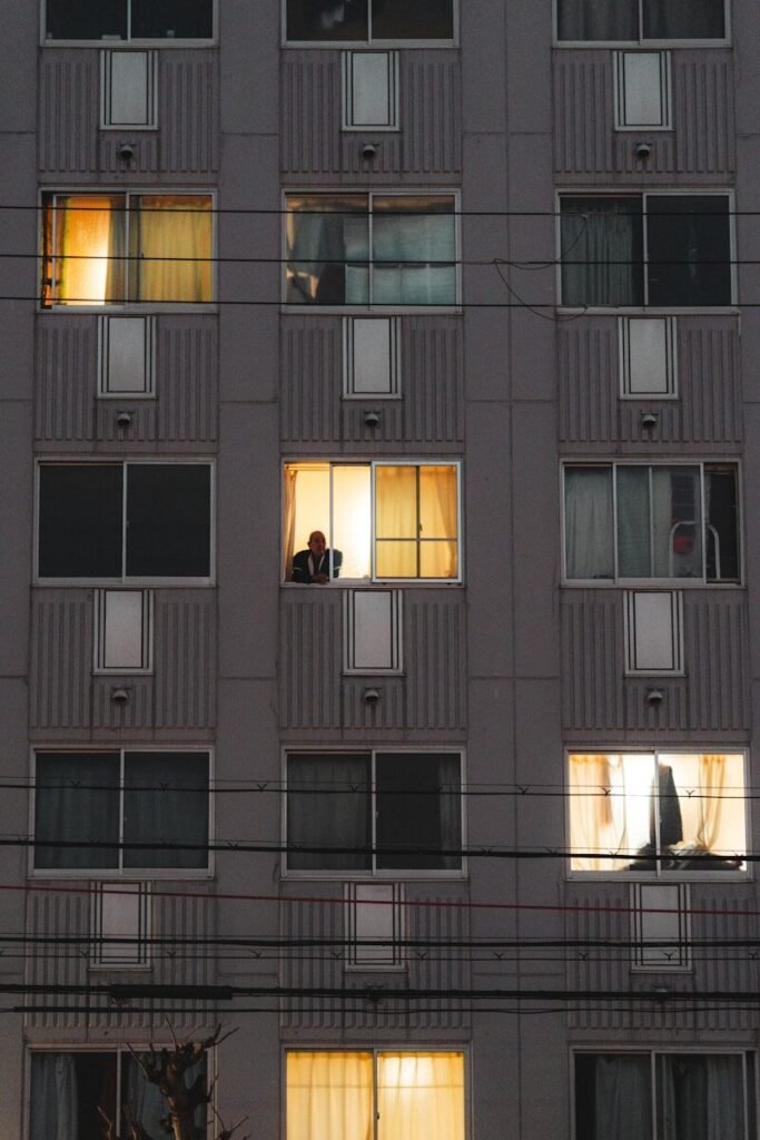 A person looks out from a brightly lit apartment window.