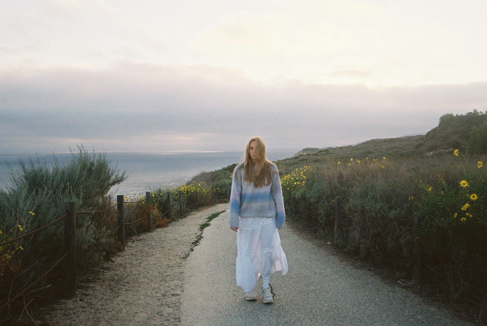 A woman walks down a coastal path at sunset.